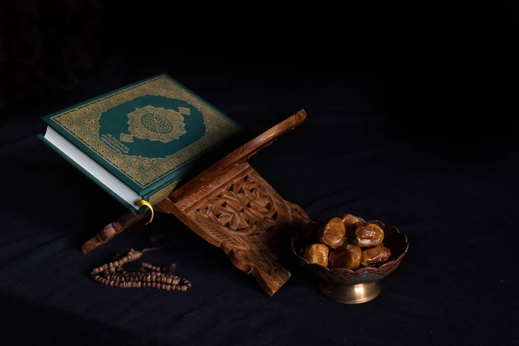 A Qur’an on a wooden stand beside dates and prayer beads on dark cloth.