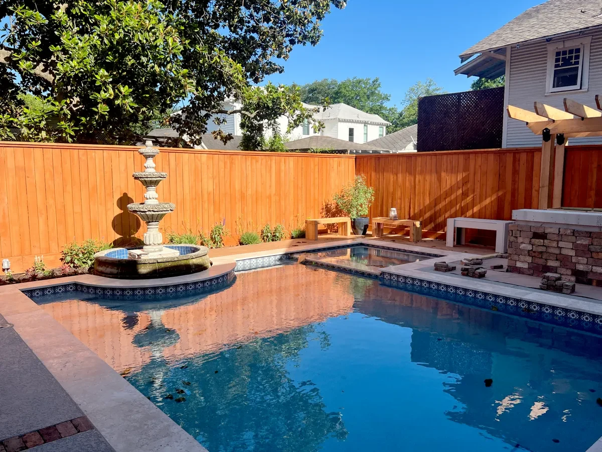 Houston Heights backyard with a tiled pool and spa, pool surround, vertical cedar fence, brick counter and pergola beside a two-story house.