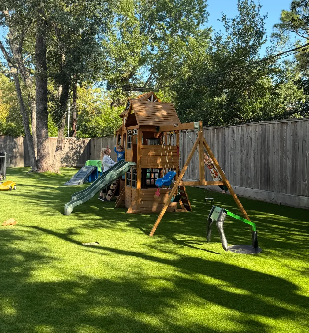 Artificial-turf play yard with wooden play equipment and a flush drainage grate beneath mature pines in Memorial, Houston.